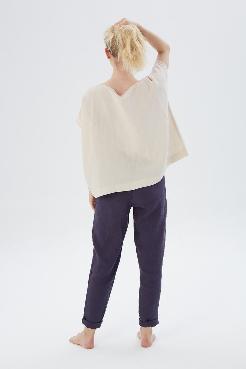 May include: A person wearing a loose-fitting, off-white linen top with a scoop neck and a pair of dark purple linen pants.