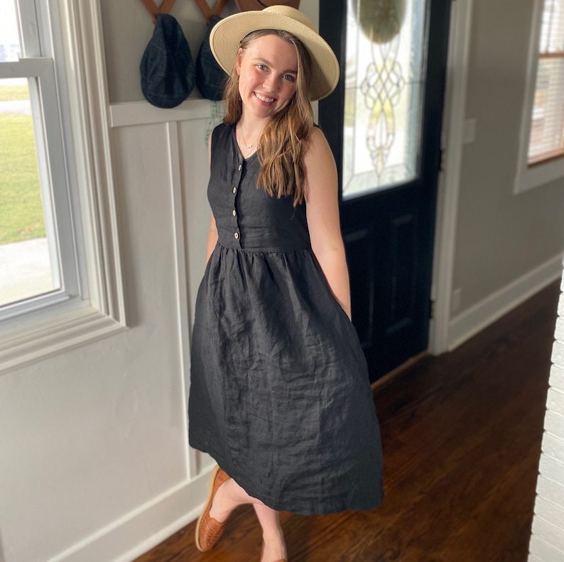 Pode incluir: Uma mulher vestindo um vestido de linho preto com bot&otilde;es na frente e um chap&eacute;u de palha. Ela est&aacute; em p&eacute; em uma porta com piso de madeira.