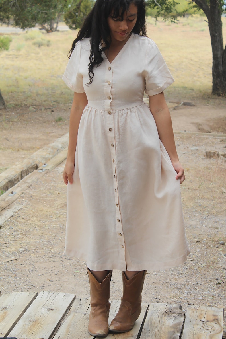 May include: A woman wearing a long, light-colored linen dress with a button-down front and short sleeves. She is wearing brown cowboy boots and standing on a wooden platform.