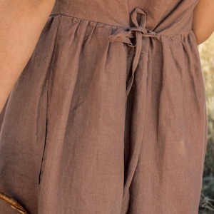 May include: Close-up of a brown linen jumpsuit. The jumpsuit features a tie at the waist and pockets. The fabric has a slightly textured appearance. The overall style is casual and comfortable.