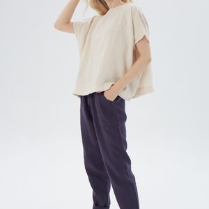 May include: A woman is wearing a loose-fitting, off-white linen top with short sleeves and a pair of dark blue linen pants. The pants have a drawstring waist and cuffed hems.