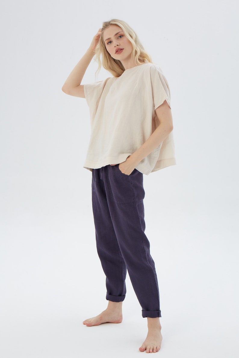 May include: A woman is wearing a loose-fitting, off-white linen top with short sleeves and a pair of dark blue linen pants. The pants have a drawstring waist and cuffed hems.