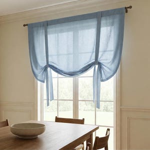 May include: A light blue tie-up curtain made of a semi-sheer fabric. The curtain is suspended from a dark metal rod and features two vertical fabric strips that are tied at the bottom to create a soft, draped, scalloped effect across the window.