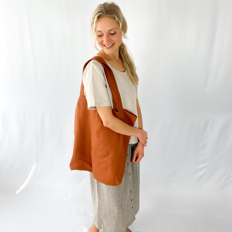 May include: A woman is wearing a light brown linen top and a striped skirt. She is carrying a large, rust-colored linen tote bag.