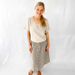 May include: A woman wearing a light beige linen top and a gray and white striped linen skirt with brown buttons down the front.