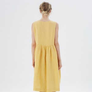 May include: A sleeveless, yellow dress with a gathered waist and a knee-length hem. The dress is made of a textured fabric and is shown from the back. The model has blonde hair styled in a braid.