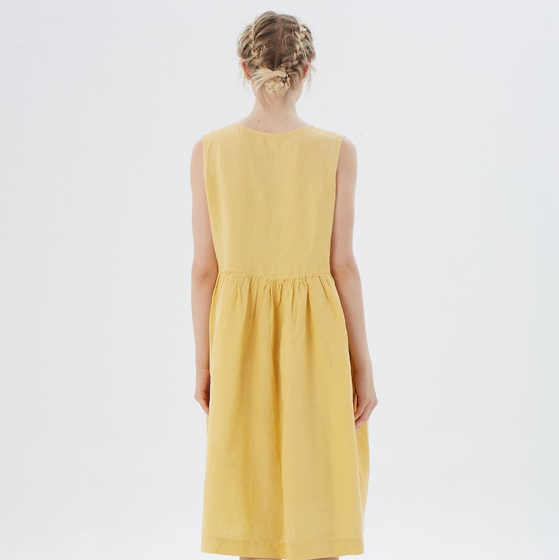 May include: A sleeveless, yellow dress with a gathered waist and a knee-length hem. The dress is made of a textured fabric and is shown from the back. The model has blonde hair styled in a braid.