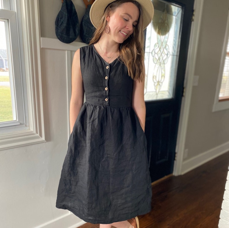 Pode incluir: Um vestido de linho preto com decote em V e fecho de bot&otilde;es. O vestido tem um corpete justo e uma saia fluida com bolsos.