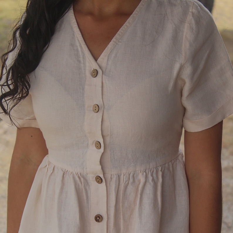 May include: A cream-colored linen dress with a v-neck and button-up front. The dress has a fitted bodice and a flowy skirt.