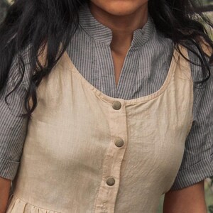 May include: A beige sleeveless dress with a button-up front and a striped shirt underneath. The dress has a relaxed fit and a simple design.