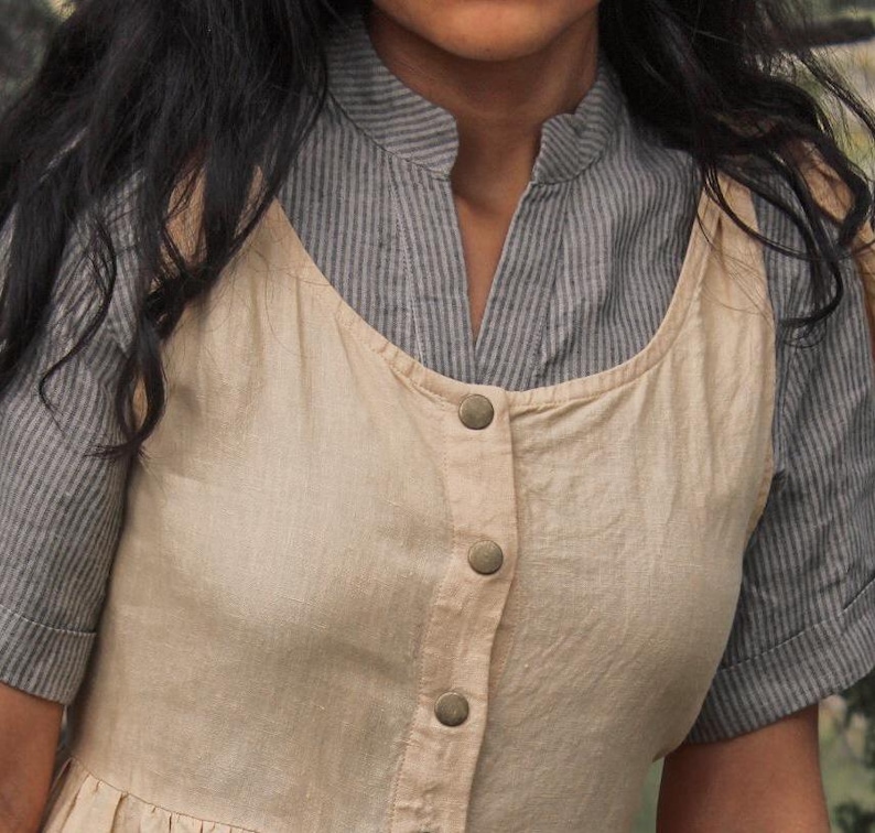 May include: A beige sleeveless dress with a button-up front and a striped shirt underneath. The dress has a relaxed fit and a simple design.