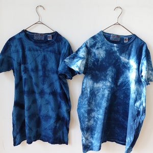 Puede incluir: Dos camisetas azules de tie-dye colgadas de perchas sobre un fondo blanco. Las camisetas tienen un patr&oacute;n de tie-dye azul claro y azul oscuro.