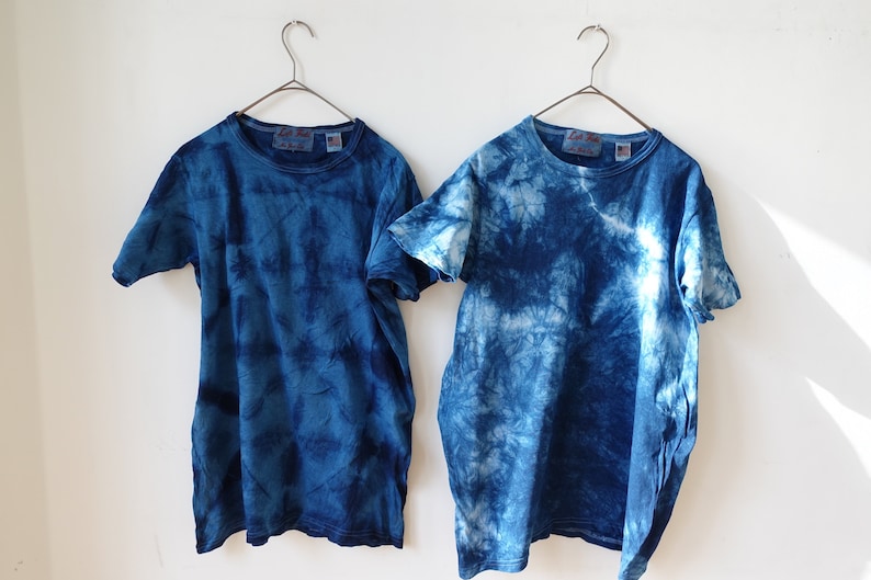 Puede incluir: Dos camisetas azules de tie-dye colgadas de perchas sobre un fondo blanco. Las camisetas tienen un patr&oacute;n de tie-dye azul claro y azul oscuro.