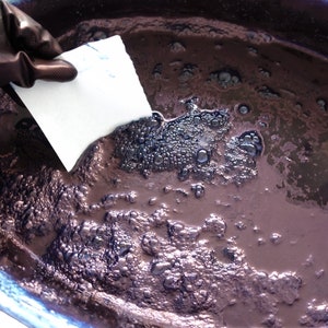Puede incluir: Primer plano de un cuenco de cer&aacute;mica blanca lleno de un l&iacute;quido morado oscuro. El l&iacute;quido tiene una consistencia espesa y viscosa y est&aacute; cubierto de peque&ntilde;as burbujas. Una mano enguantada sostiene una toalla de papel blanca sobre el l&iacute;quido.