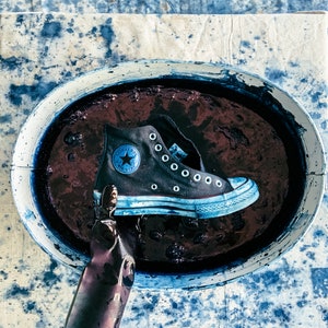Puede incluir: Una zapatilla Converse azul y blanca est&aacute; sumergida en un ba&ntilde;o de tinte azul. La zapatilla tiene un logotipo de estrella negra y el texto "Converse" en el lateral.