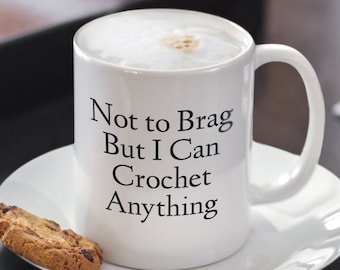 May include: White ceramic coffee mug with a black inscription that reads "Not to Brag But I Can Crochet Anything". The mug is filled with coffee and sitting on a white saucer with a cookie on the side.