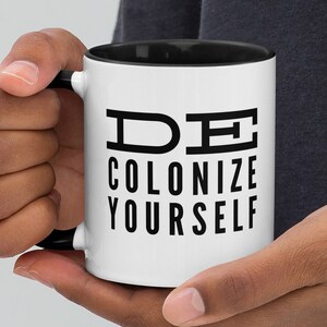 Könnte beinhalten: Eine weiße Keramiktasse mit schwarzem Rand und Henkel. Die Tasse hat einen schwarzen Text mit der Aufschrift "DE COLONIZE YOURSELF".