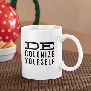 Könnte beinhalten: Weiße Keramiktasse mit schwarzem Text, der "DE COLONIZE YOURSELF" lautet.