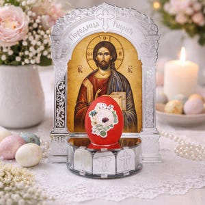 May include: A decorative Easter display featuring a red egg with a floral sticker, a religious icon, and a lit candle. The scene includes pastel-colored eggs, flowers, and a string of pearls, all set on a white lace tablecloth.