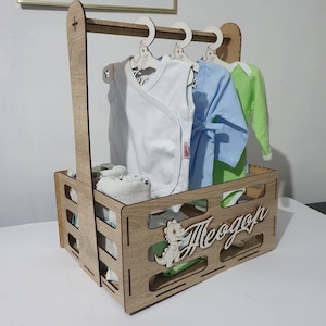 May include: A wooden baby clothes organizer with a handle and a name cutout. The organizer holds baby clothes on hangers and folded items. The clothes are white, blue, and green. A small dinosaur is cut out on the side.