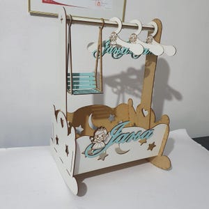 Könnte beinhalten: Ein hölzernes Baby-Schaukel- und Kleiderbügel-Set. Die Schaukel und die Kleiderbügel sind hellblau und weiß. Die Schaukel hat einen kleinen Sitz. Die Kleiderbügel haben ein Prinzessinnen-Design. Der Name "Jansa" steht auf der Vorderseite der Schaukel.