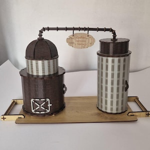 以下が含まれることがあります： 2 つの円筒形の構造がパイプで接続された木製の蒸留所の模型。大きな構造は茶色でドアがあり、小さな構造は縞模様です。それらの間に看板がぶら下がっています。