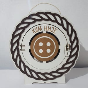 May include: A decorative wooden plaque with a circular design. The plaque features a brown rope-like border, a white interior with a brown button design, and the text "KUM NIJE". It has a stand and a rope for hanging.