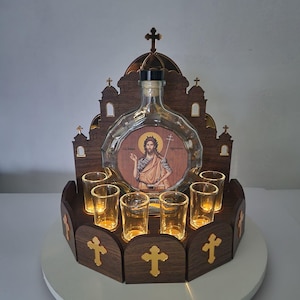 Peut inclure: Un ensemble de service à liqueur décoratif en bois avec une bouteille en verre transparent représentant une image d'une figure religieuse. L'ensemble comprend des verres à shot et est éclairé par un éclairage chaleureux. La base en bois est ornée de croix.