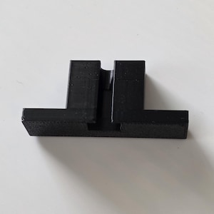 May include: A black, 3D-printed phone stand. The stand has a rectangular base with two vertical supports and a slot to hold a phone. The stand is made of a matte black material.