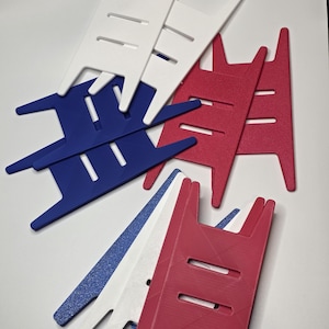 May include: A collection of colorful, plastic yarn swift tools in red, white, blue, and glittery blue. These tools have a unique shape with a central bar and angled arms, designed for holding yarn skeins. The tools are arranged on a white surface.