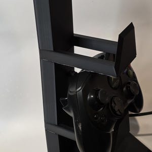 May include: A black, geometric stand designed to hold a gaming controller. The stand has a tall, rectangular back with a horizontal support and a base. An Xbox controller is placed in the stand.