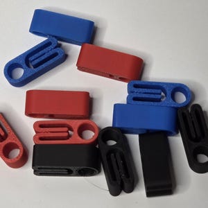 May include: Assortment of 3D-printed plastic clips in various colors, including red, blue, and black. The clips have a rectangular shape with a slot and a circular hole, designed for securing or fastening.