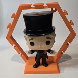 May include: A Funko Pop figure of a man in a black suit and top hat, with a red tie, displayed in an orange hexagonal stand. The figure has a light skin tone and gray hair. The stand has a base and a frame.