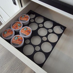 May include: A drawer with a black organizer holding several Starbucks Pumpkin Spice coffee pods. The drawer is part of a white cabinet with a dark brown accent and a sliding door mechanism.
