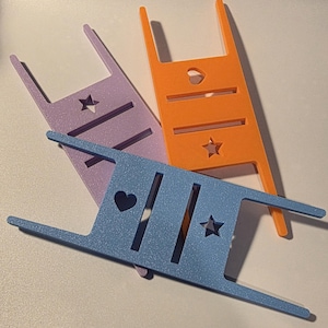 May include: Three colorful yarn swift tools in lavender, orange, and blue. Each tool has a rectangular body with two parallel slots and a decorative cutout of a heart or star. The tools have two vertical posts and two horizontal arms.