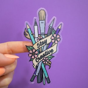 May include: A clear sticker with a design of various art supplies, including a paintbrush, pencils, and a pen, crossed over each other. The supplies are tied together with a banner that reads "Stay Creative".