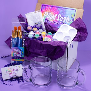 May include: A white box with a purple tissue paper lining, filled with a paint your own mug kit. The kit includes two clear mugs, paint, brushes, a paint palette, and a thank you card. The box also has a sticker that says "This product plants one tree."