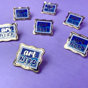 以下が含まれることがあります： 青と白のデザインの金色のエナメルピン。ピンには波打つフォントで「art nerd」という文字が書かれています。