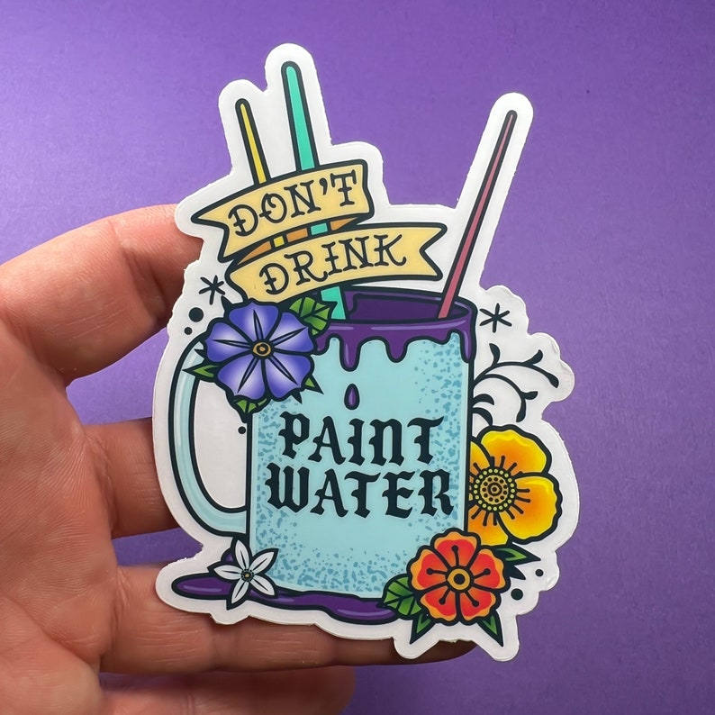 Op de afbeelding: Een blauwe mok met de tekst "Drink geen verfwater" en een paarse rand. De mok is omringd door bloemen en kwasten.
