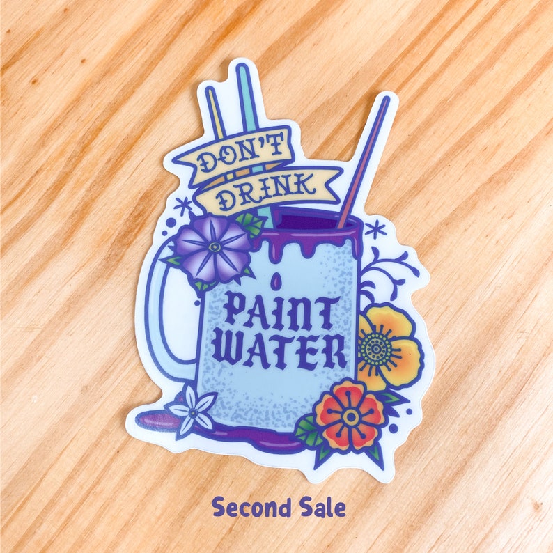 Op de afbeelding: Een blauwe en paarse mok met de tekst "Drink geen verfwater" en twee rietjes die uit de bovenkant steken. De mok is versierd met bloemen en wijnstokken.