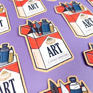 K&ouml;nnte beinhalten: Aufkleber mit einem Zigarettenschachtel-Design, die das Wort "ART" und Kunstutensilien zeigen. Die Aufkleber haben ein rot-wei&szlig;es Farbschema mit dem Text "MAKE SOME ART" und "HIGHLY ADDICTIVE".