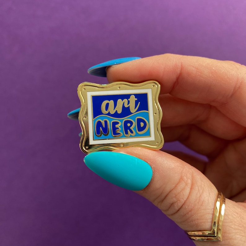 以下が含まれることがあります： 青と白のデザインの金色のエナメルピン。ピンには「art nerd」という文字がスタイリッシュなフォントで書かれています。