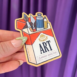 K&ouml;nnte beinhalten: Ein Aufkleber in Form einer Zigarettenschachtel mit Kunstutensilien. Die Schachtel ist rot und wei&szlig;, mit den Worten "MAKE SOME ART" und "HIGHLY ADDICTIVE". Im Inneren befinden sich ein Pinsel, ein Spachtel, eine Farbtube und ein Bleistift.
