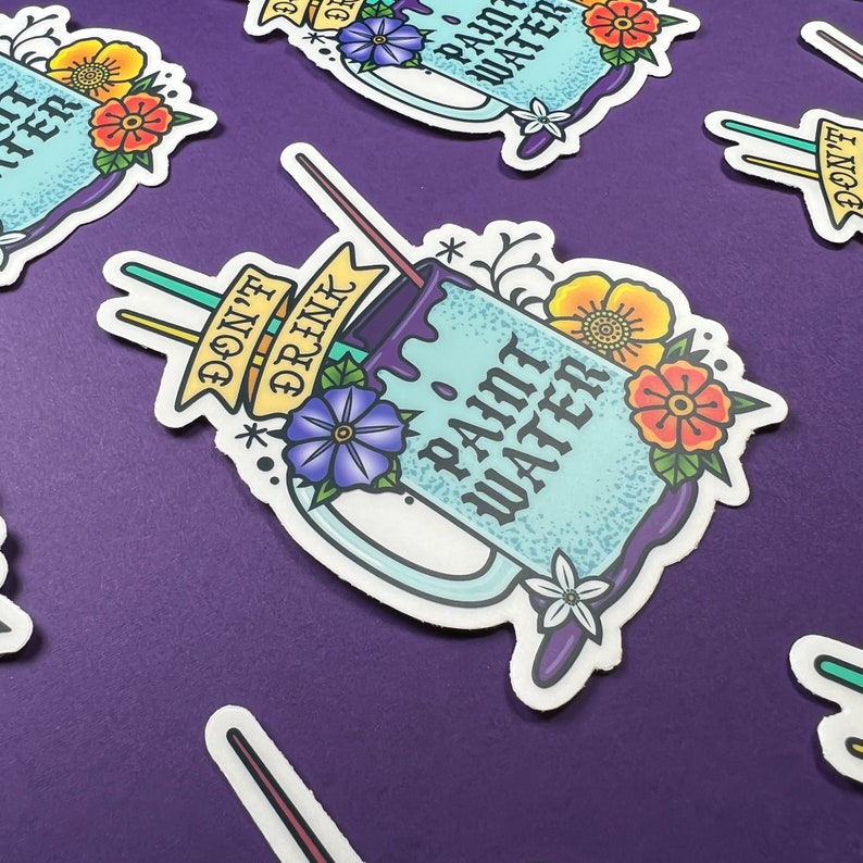 Op de afbeelding: Een blauwe en witte mok met de tekst "Drink geen verfwater" en een paarse druppel. De mok is versierd met bloemen en twee rietjes.