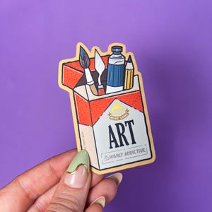 K&ouml;nnte beinhalten: Ein Aufkleber mit einer Cartoon-Illustration eines Zigarettenp&auml;ckchens mit Kunstutensilien. Das P&auml;ckchen ist rot und wei&szlig;, mit dem Wort "ART" und dem Satz "HIGHLY ADDICTIVE" darauf. Darin befinden sich ein Pinsel, ein Spachtel, eine Farbtube und ein Bleistift.