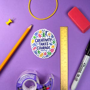 May include: A circular sticker with a white background featuring the text 'CREATIVITY TAKES COURAGE -MATISSE' in dark blue, surrounded by colorful abstract leaf and organic shapes in red, yellow, green, and blue, next to a 6-inch metal ruler.
