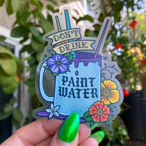 Op de afbeelding: Een blauwe en witte mok met de tekst "Drink geen verfwater" en bloemmotieven. De mok is omringd door kwasten en een palet.