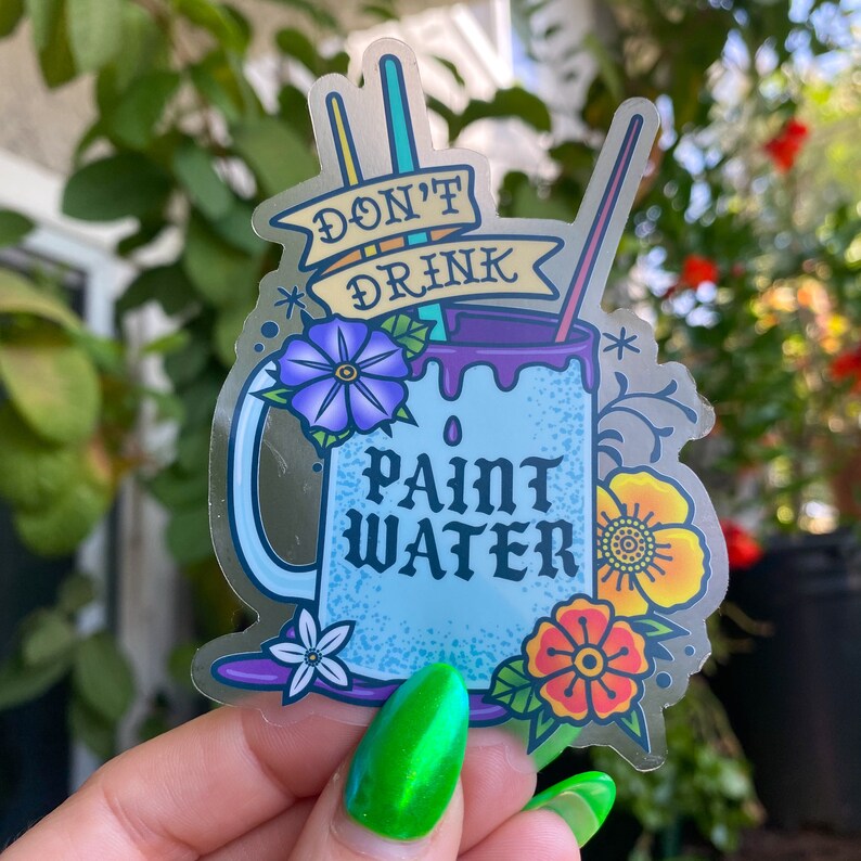Op de afbeelding: Een blauwe en witte mok met de tekst "Drink geen verfwater" en bloemmotieven. De mok is omringd door kwasten en een palet.