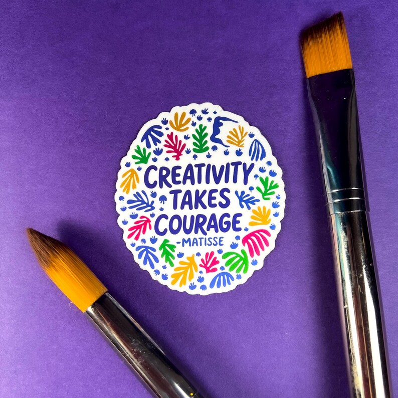 May include: A round white sticker featuring the blue text "CREATIVITY TAKES COURAGE -MATISSE" surrounded by a colorful pattern of abstract organic shapes in red, green, yellow, and blue, positioned between two paintbrushes with orange bristles.