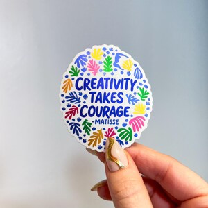 May include: A circular sticker with a scalloped edge featuring the blue text "CREATIVITY TAKES COURAGE -MATISSE" in the center. The design is surrounded by a colorful pattern of abstract organic leaf-like shapes in yellow, pink, green, and blue.